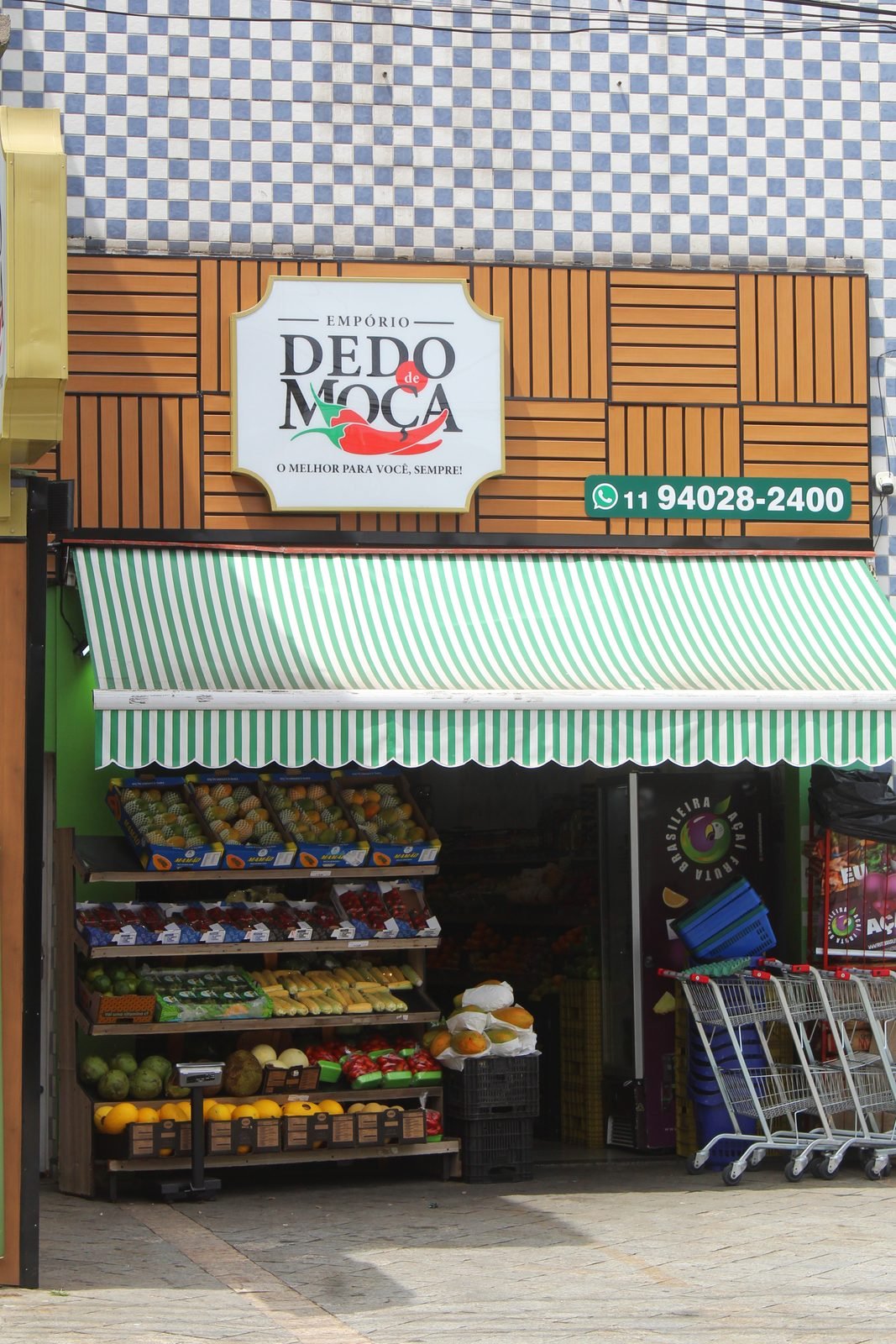 Frente do Empório Dedo de Moça com a placa, o toldo listrado e as bancas de frutas na entrada
